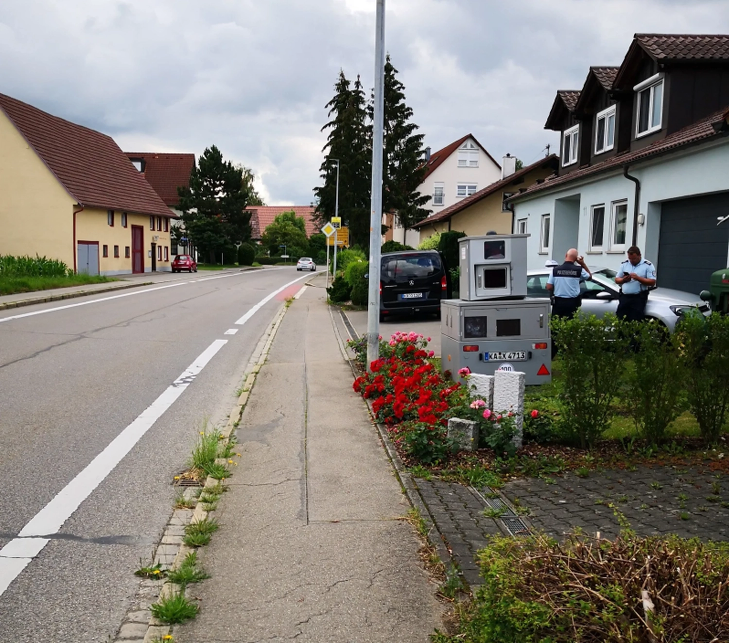 Radarrent GmbH - Verkehrsüberwachung - Zwei Polizisten stehen in der Nähe einer Geschwindigkeitskamera in einer Wohnstraße mit Häusern und roten Blumen am Gehweg.