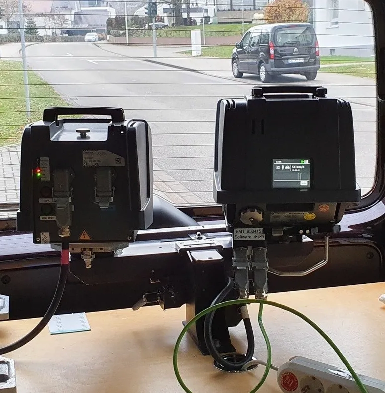 Radarrent GmbH – Verkehrsüberwachung – Zwei elektronische Geräte sind über Kabel mit dem Armaturenbrett eines Fahrzeugs verbunden. Auf dem rechten Gerät wird auf einem kleinen Bildschirm Geschwindigkeit und Zeit angezeigt