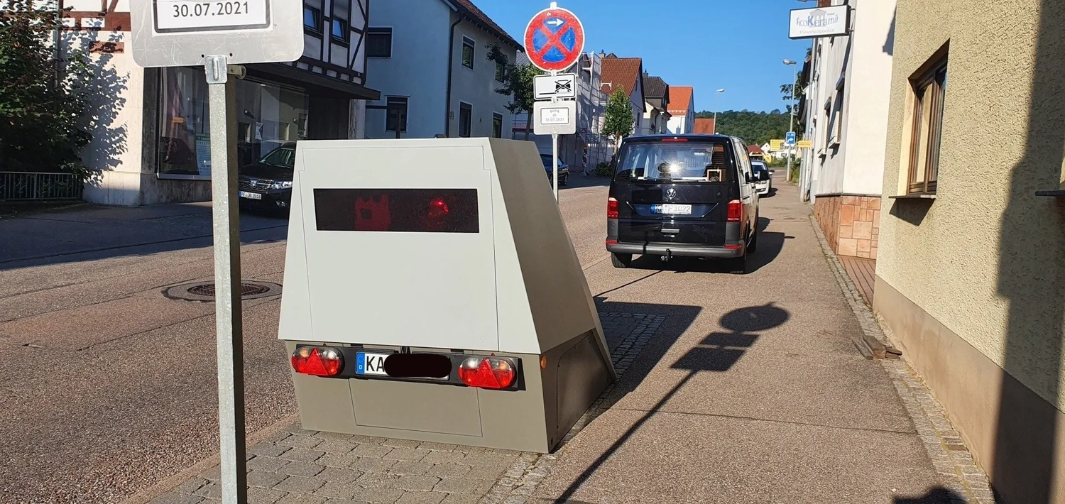 A speed camera device installed on a sidewalk next to a no parking sign, with cars parked along a street in a residential area.