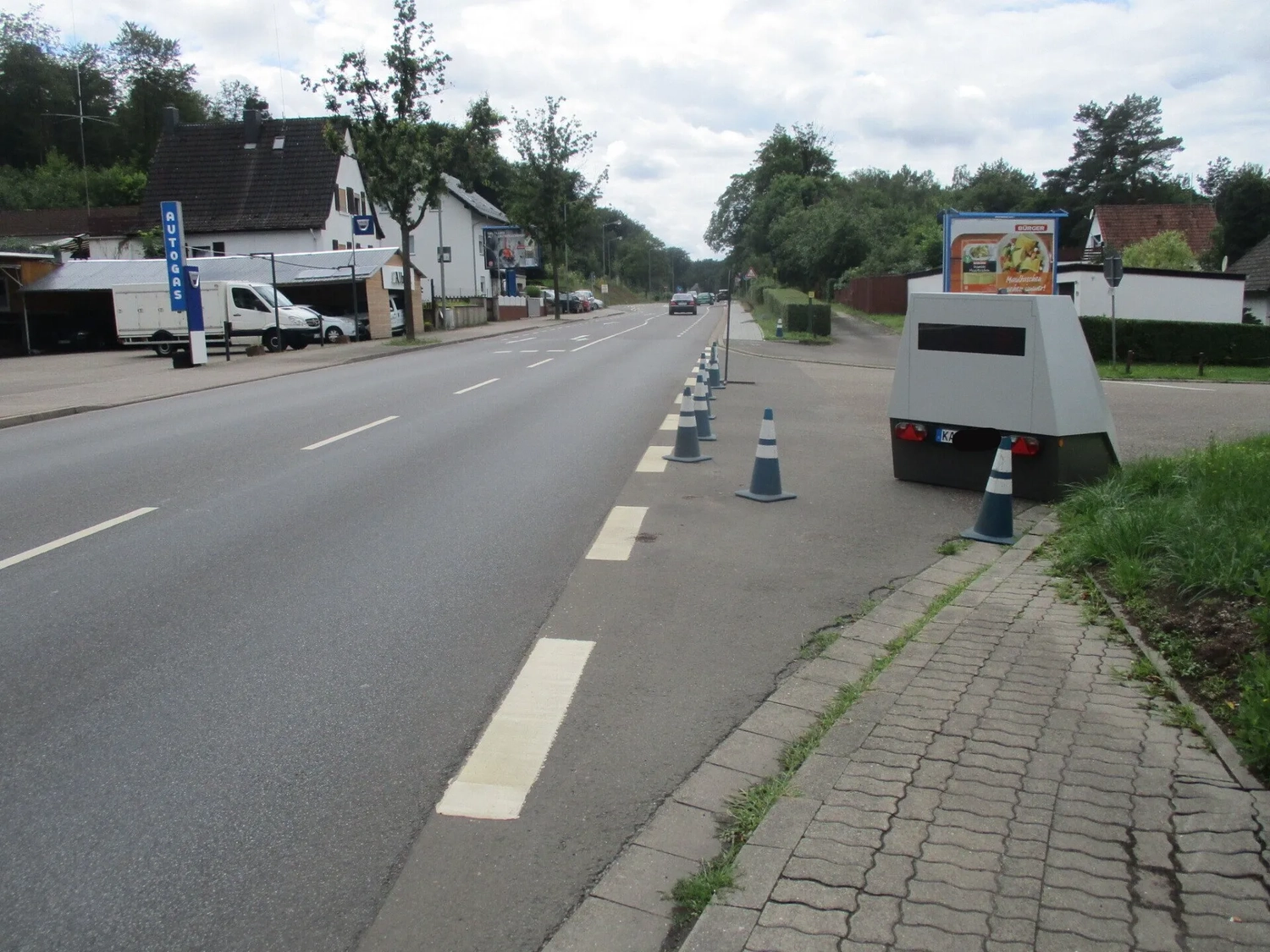 Radarrent GmbH – Verkehrsüberwachung – Leere zweispurige Straße mit blauen Verkehrskegeln am Gehweg und einem grauen Blitzer auf der rechten Seite unter bewölktem Himmel