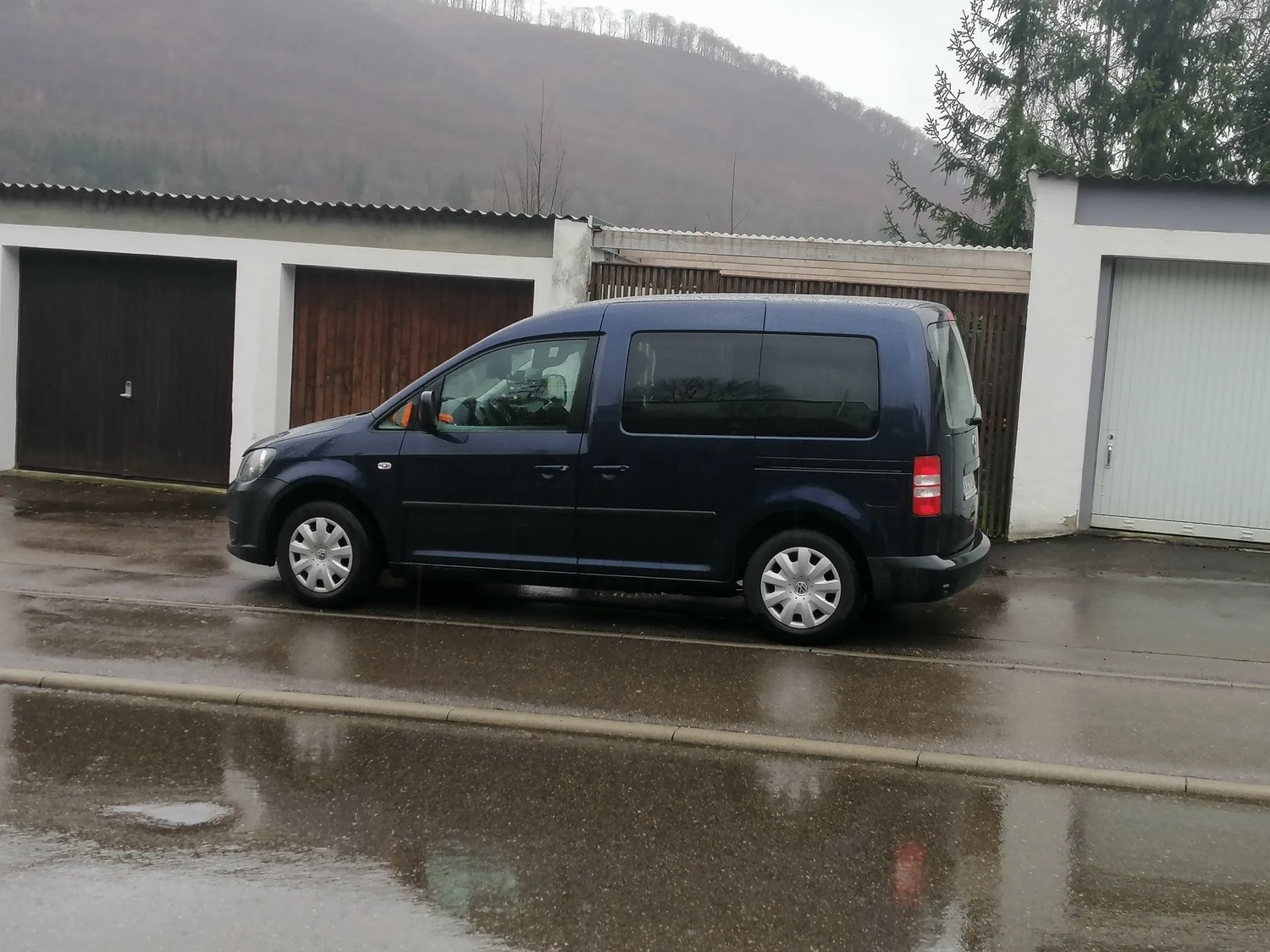 Radarrent GmbH – Verkehrsüberwachung – Blauer VW-Transporter parkt auf einer nassen Straße vor geschlossenen Garagen; im Hintergrund erhebt sich ein Hügel.