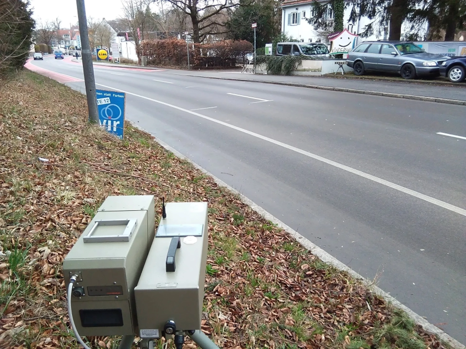 Radarrent GmbH – Verkehrsüberwachung – Graues Straßenüberwachungsgerät auf einem Stativ in der Nähe einer Straße mit Autos und Gebäuden im Hintergrund