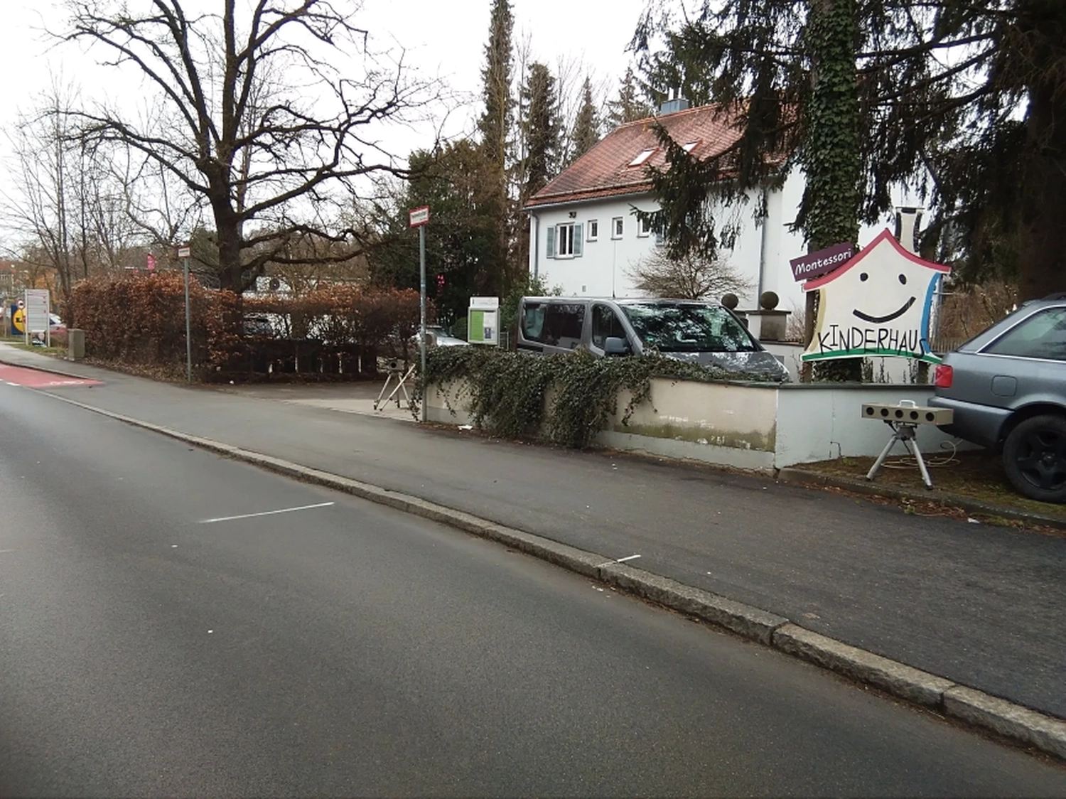 Radarrent GmbH - Verkehrsüberwachung - Straßenansicht eines Gehwegs mit geparkten Autos und einem farbenfrohen Montessori-Kinderhaus-Schild in Form eines lächelnden Hauses neben einem weißen Gebäude.