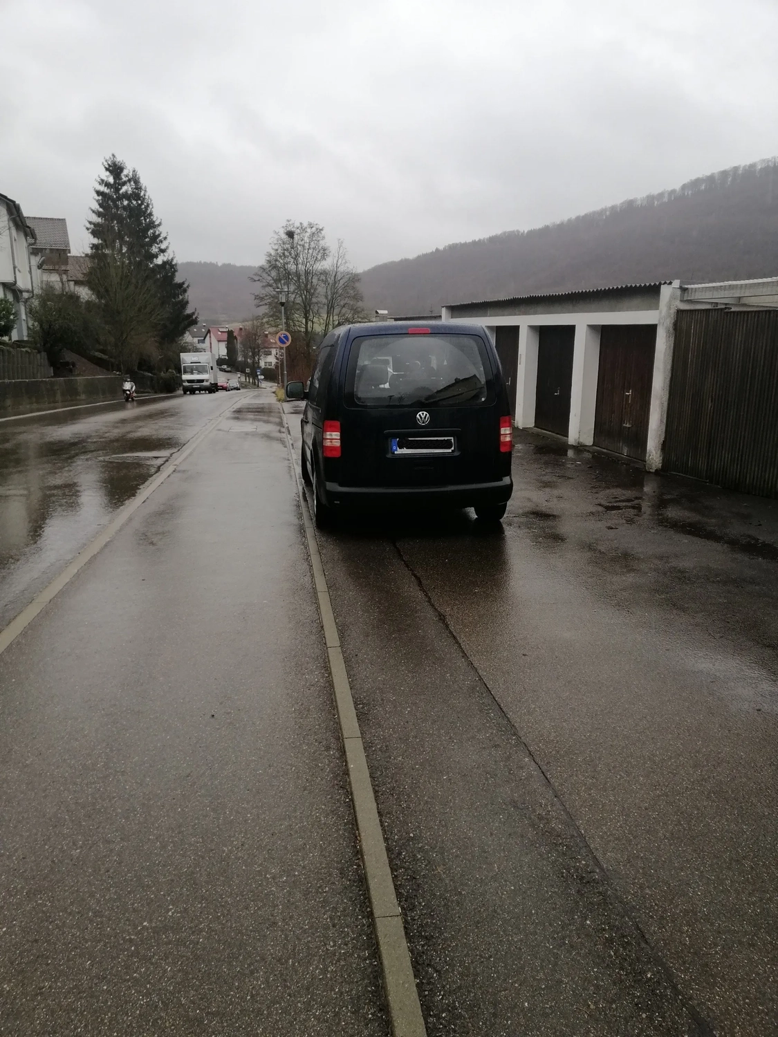 Radarrent GmbH – Verkehrsüberwachung – Schwarzer Volkswagen-Minivan parkt an einem bewölkten Tag auf einer nassen Straße neben Garagen.