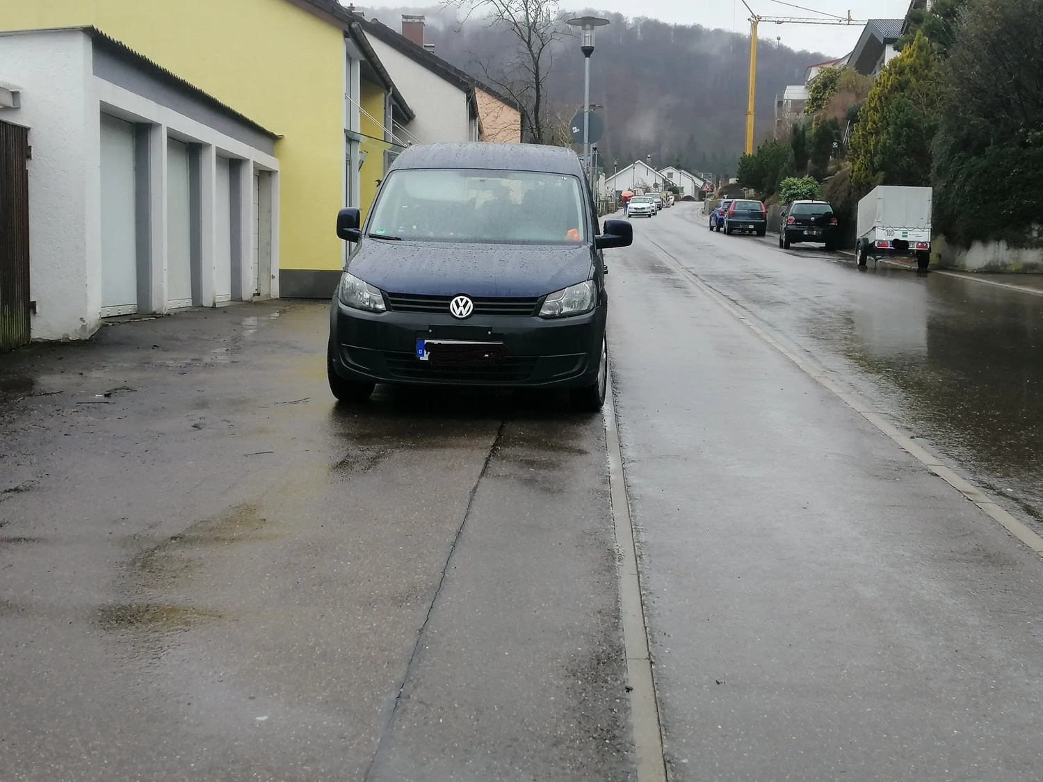 Radarrent GmbH – Verkehrsüberwachung – Vorderansicht eines nassen VW-Transporters, der auf einer regnerischen Straße in der Nähe von Wohnhäusern und einem Hügel im Hintergrund geparkt ist.