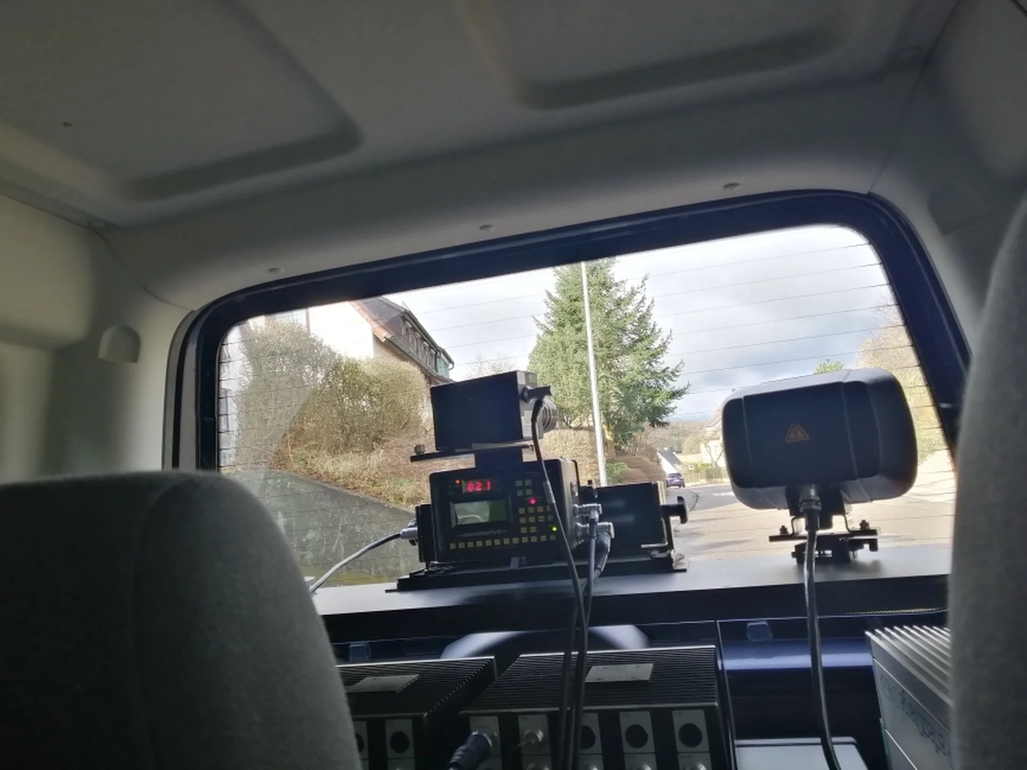 Radarrent GmbH – Verkehrsüberwachung – Blick durch die Heckscheibe eines Autos auf montierte elektronische Geräte und eine Straße mit Bäumen und Häusern im Hintergrund