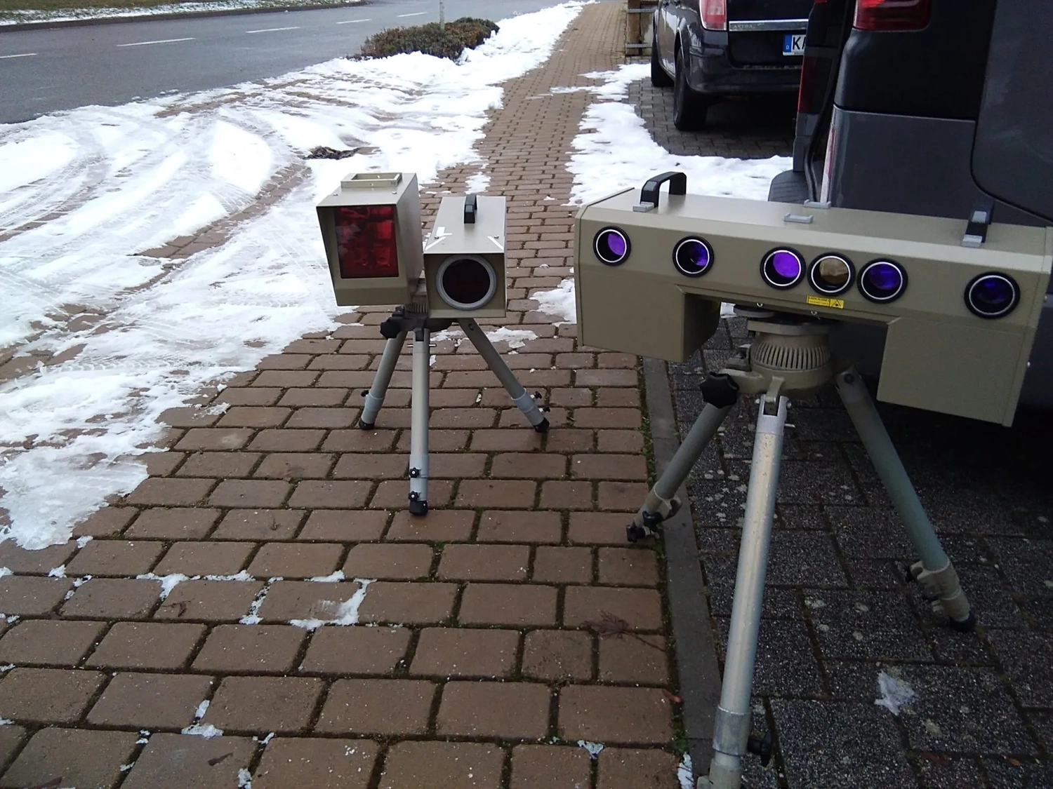 Two tripod-mounted speed radar cameras positioned on a snow-dusted sidewalk beside parked cars.
