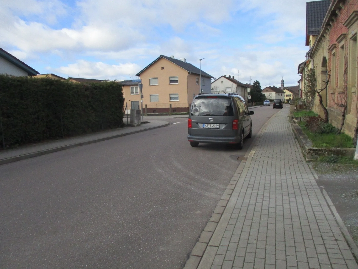 Radarrent GmbH – Verkehrsüberwachung – Ein grauer VW-Minivan parkt in einer Wohnstraße mit Häusern, einem Bürgersteig und einer Hecke unter einem teilweise bewölkten Himmel