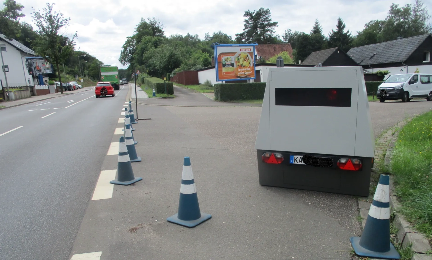 Radarrent GmbH – Verkehrsüberwachung – Mobile Geschwindigkeitsmessanlage hinter Verkehrskegeln am Straßenrand mit Autos und Häusern im Hintergrund
