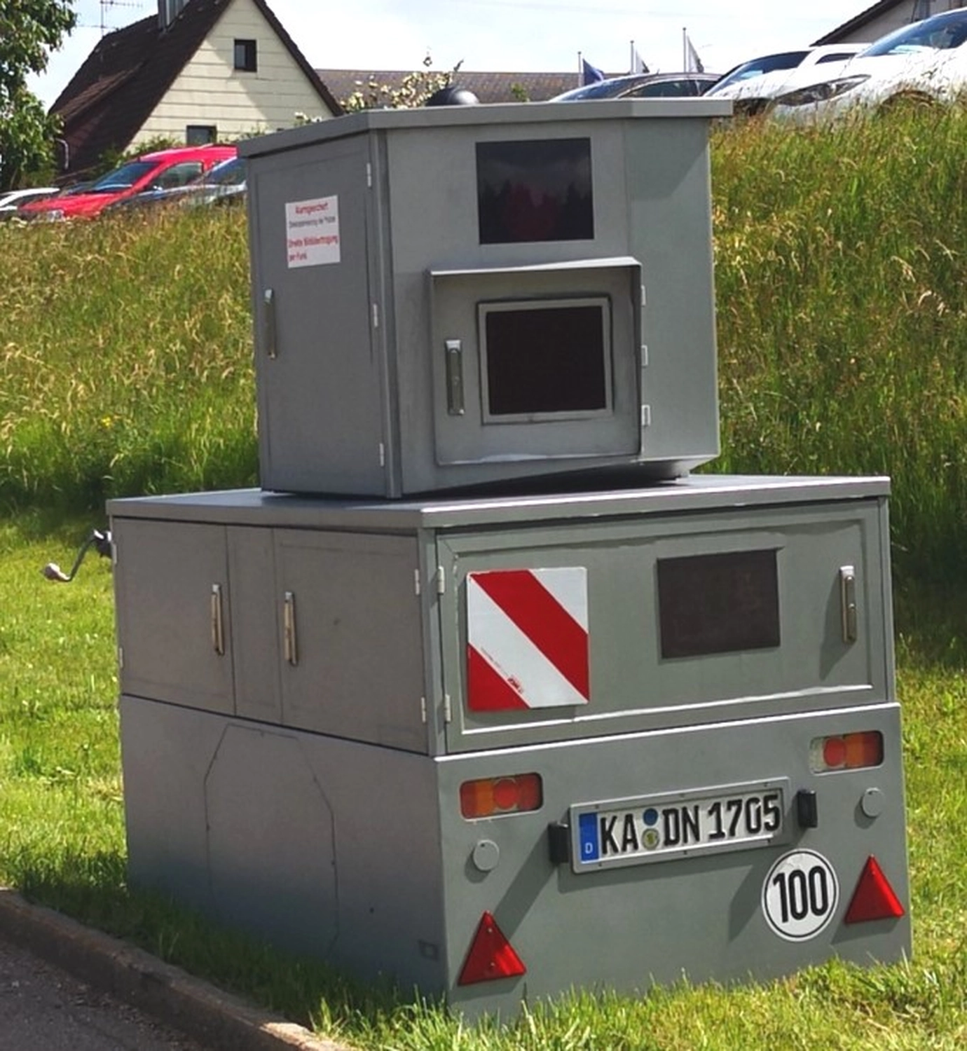 Gray roadside speed camera with a license plate and traffic signs on a grassy area next to a road.