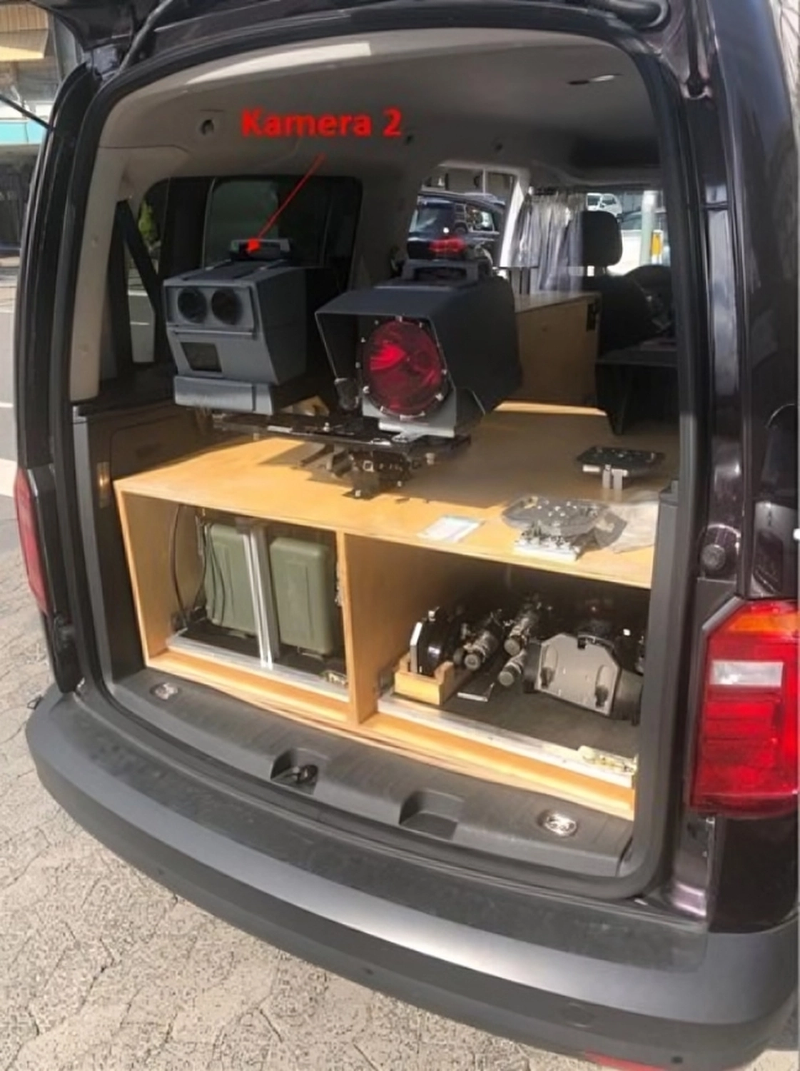Open rear of a black vehicle equipped with mounted radar and camera devices labeled 'Kamera 2' inside a custom wooden enclosure.